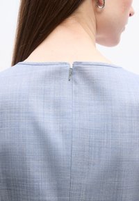 Πλάτη ενός ατόμου που φοράει ένα ανοιχτό μπλε υφασμάτινο μπλουζάκι με ένα μικρό ασημένιο φερμουάρ στο λαιμό και ένα ασημένιο κρεμαστό σκουλαρίκι.