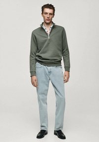 Felpa verde a zip con colletto a coste, abbinata a jeans azzurri. Il modello è in posizione eretta, scarpe nere visibili, sfondo neutro.