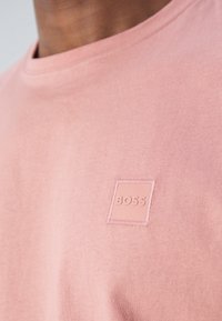 Gros plan sur un t-shirt rose à col rond avec un petit patch carré portant le mot "BOSS" sur la poitrine.