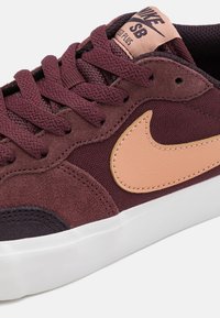 Tenis burgund cu piele nubuc, cu un finisaj texturat, având un logo Nike de culoare piersică deschisă, vârf rotunjit și talpă din cauciuc alb.