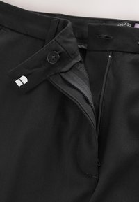 Pantalon noir avec fermeture éclair ouverte et bouton, montrant la taille et la texture du tissu.