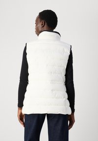 Vit pufferväst med hög krage och vertikal sömnad, tillsammans med en svart långärmad topp och mörka jeans. Slät textur, ingen synlig hårdvara.