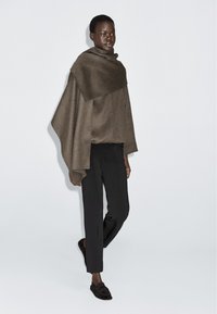 Woman wearing a brown wool cape over black trousers and dark loafers, standing against a plain light gray background.