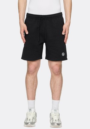 Mann trägt schwarze Shorts mit Kordelzug und Logo am Bein, weiße Socken und weiße Sportsneaker und steht vor einem einfarbigen Hintergrund.