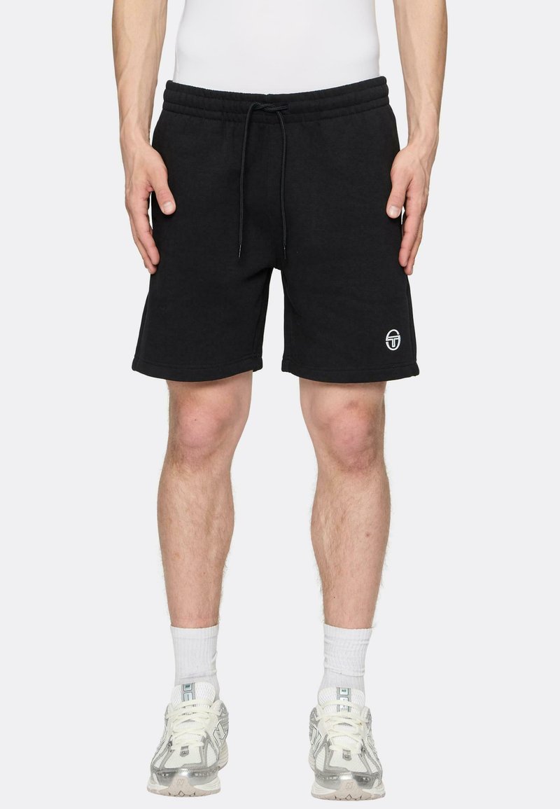 Mann som har på seg svarte shorts med snor og logo på benet, hvite sokker og hvite sportsko, står mot en ensfarget bakgrunn.