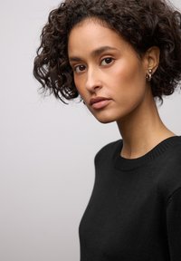 Pull noir à encolure ronde avec des détails côtelés, porté par une personne aux cheveux bouclés, sur un fond neutre.