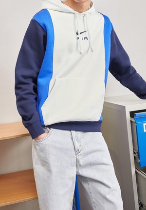 Personne portant un sweat à capuche Nike blanc et bleu et un jean délavé clair, debout avec une main dans la poche à côté d'un classeur et d'une étagère en bois.