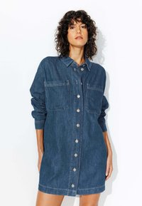 Robe chemise en denim bleu foncé avec des manches longues, un devant boutonné, deux poches poitrine et une coupe décontractée. Texture lisse et design classique.