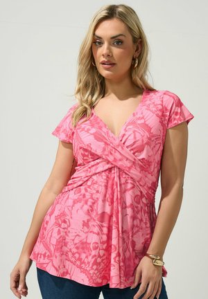 Blondynka w różowej bluzce z krótkim rękawem w motyle, z złotymi kolczykami-kołami i bransoletkami, pozująca na jednolitym tle.