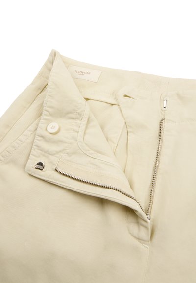 Pantalones beige con cremallera parcialmente abierta, botón y cierre de gancho y barra, mostrando la pretina doblada y la etiqueta interior que dice "SLOWEAR".