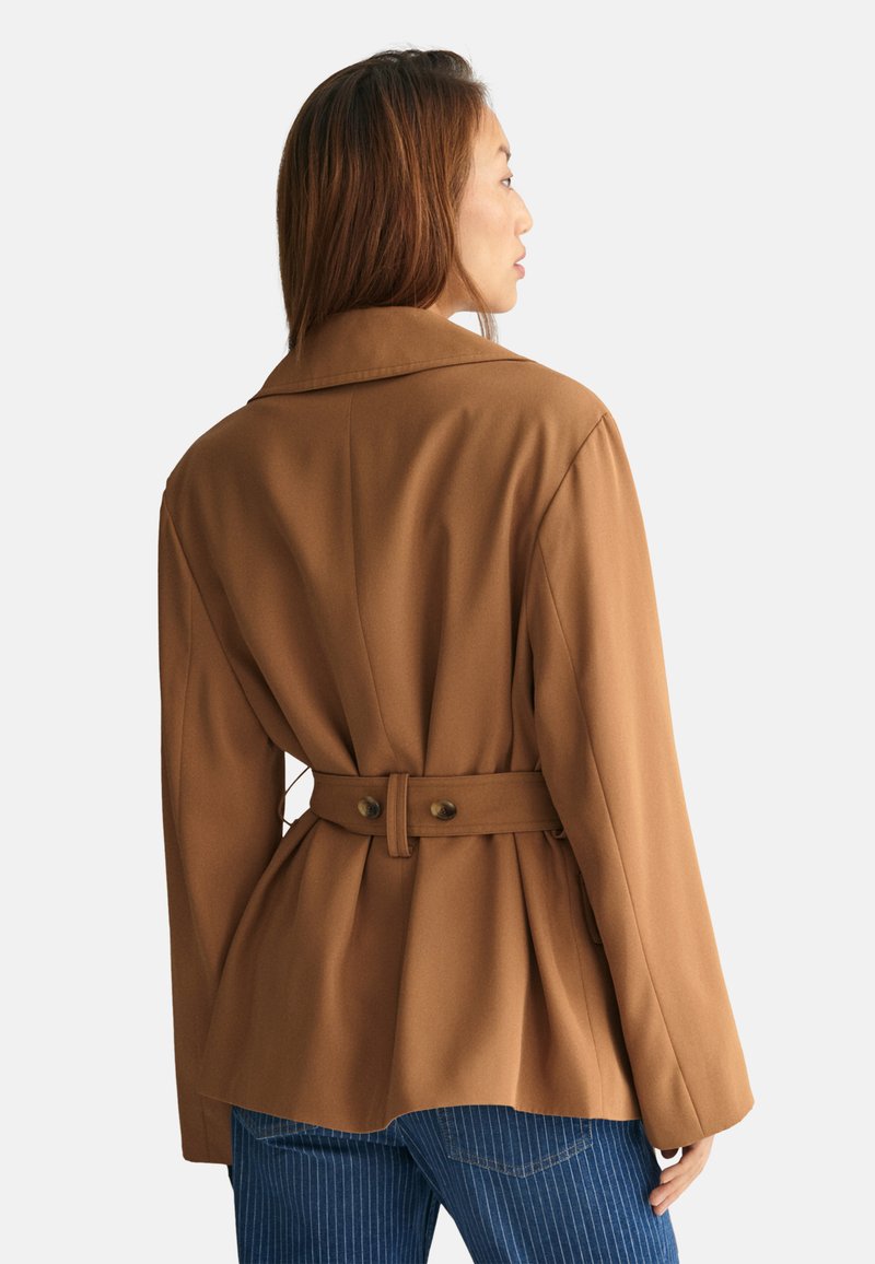 Veste marron ceinturée avec un large col, coupe ajustée et dos plissé. Dotée de deux boutons à la taille. Portée sur un jean bleu à rayures.