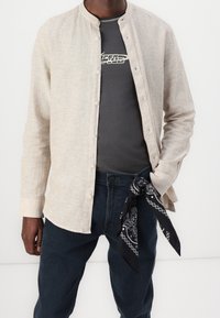 Camisa beige de manga larga con botones y cuello mandarín sobre una camiseta gris con diseño gráfico. Pantalones vaqueros negros con un pañuelo negro atado a la cintura.