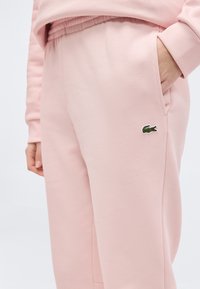 Άτομο που φοράει ανοιχτό ροζ παντελόνι φόρμας με ελαστική μέση, χέρια στις τσέπες και μικρό πράσινο λογ ότυπο κροκόδειλου στον μηρό.