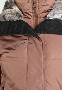 Chaqueta acolchada marrón con un cuello alto, con una superposición de fieltro negro y un ribete de piel gris clara. Cierre central con cremallera y detalles de costura vertical.