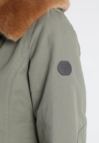 Olive zelená bunda s kožešinovým límcem, která má texturovanou tkaninu, detaily na švech ramen a kulatou černou nášivku s logem na rukávu.