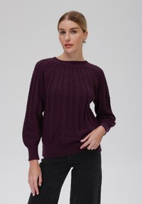 Lila gestrickter Pullover mit einem strukturierten Design, featuring einem Kabelstrickmuster und gerippten Bündchen, kombiniert mit schwarzen hochgeschnittenen Hosen.