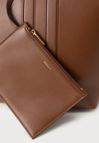 Καφέ δερμάτινη τσάντα χειρός με υφή στην επιφάνεια, συνοδευόμενη από μια μικρότερη τσαντούλα σε ίδια απόχρωση. Χρυσοί φερμουάρ και λογότυπο στην τσαντούλα. Καθαρές γραμμές και μινιμαλιστικός σχεδιασμός.