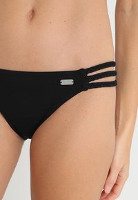 Bas de bikini noir avec trois bretelles tressées de chaque côté et un accent en métal argenté. Tissu lisse avec un design classique.