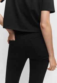 Henkilö, jolla on päällä mustat korkeavyötäröiset pillifarkut, joissa on takataskut, ja käsi asetettu vasempaan taskuun, yhdistettynä mustaan lyhyeen toppiin.