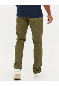Pantalons en mélange de coton olive avec une coupe slim, deux poches arrière et ourlets roulés. Associés à des baskets blanches et une chemise bleue.