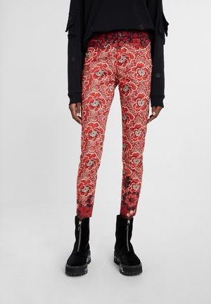 Person trägt rote Blumenmuster-Hose, schwarzes langärmeliges Oberteil und schwarze Stiefeletten mit vorderen Reißverschlüssen und bedruckten Sohlen.