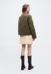 Woman stands facing sideways wearing an oversized olive green jacket, beige skirt, and black knee-high boots on a plain white background.