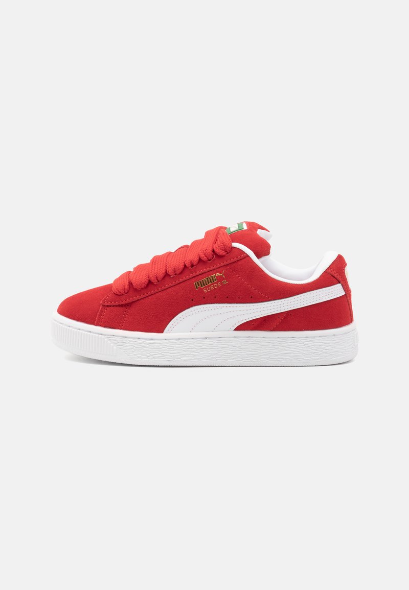 Baskets en suede rouge avec des accents blancs, semelle texturée et lacets rouges épais. Présentent un logo doré sur le côté et une étiquette verte et blanche sur la languette.