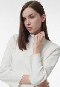 Witte, lange mouwen gebreide coltrui met een zachte textuur. Model poseertdenken met de armen gekruist, hand bij de kin.