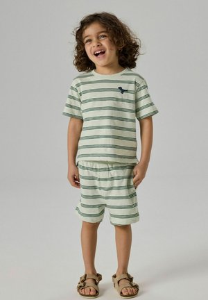 Μικρό παιδί με σγουρά μαλλιά που χαμογελάει, φοράει μπλουζάκι με μπεζ και πράσινες ρίγες και κοντά παντελονάκια σετ με ταμπά σανδάλια.