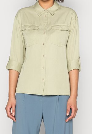 Personne portant une chemise vert clair boutonnée avec deux poches poitrine et des manches retroussées, associée à un pantalon bleu ample.
