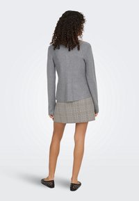Femme aux cheveux bouclés portant un haut gris à manches longues, une mini-jupe à carreaux et des ballerines noires cloutées, debout dos au spectateur.