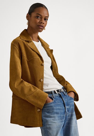 Nő barna színű, gombos velúrdzsekit, fehér bordázott inget, fekete övvel ellátott kék farmert visel, zsebében tartja a kezét, és a kamerába néz.