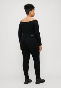 Kvinne med krøllete hår iført et svart av-shoulder topp, svarte jeans og svarte ankelstøvler, stående mot en enkel, lys bakgrunn.