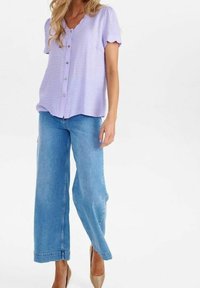 Donna in blusa button-down a maniche corte color lavanda con maniche scallop, jeans a gamba larga blu e tacchi beige, in piedi su uno sfondo bianco.