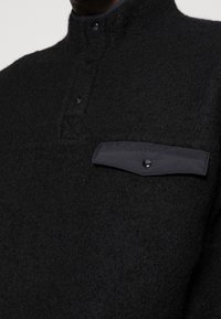 Mustapaita, jossa on teksturoitu villasekoite, korkea kaulus, napitus ja erottuva musta läpätasku, jossa on napin yksityiskohta.