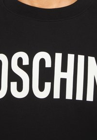 Detailní záběr na černou tkaninu s velkými bílými písmeny tvořícími část názvu značky na oblečení poblíž výstřihu.