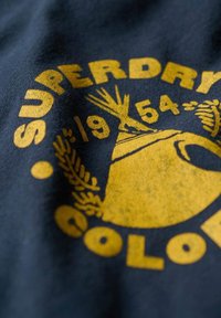 Tela azul oscura con un logo impreso en amarillo llamativo que incluye texto, ilustraciones y detalles que asemejan un sombrero y hojas. La textura es suave.