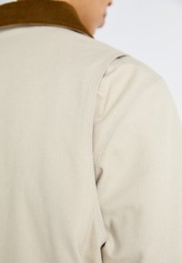 Prenda de algodón beige con un cuello de pana marrón. Primer plano en el área del hombro y el cuello, destacando las costuras y la textura.
