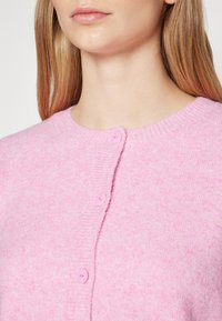 Ljusrosa kofta med ribbat halsringning, fem rosa knappar framtill och ett mjukt, texturerat tyg.
