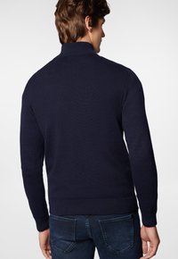 Granatowy sweter dzianinowy z wysokim kołnierzem i ściągaczami, charakteryzujący się fakturowanym materiałem i długimi rękawami. Widok z tyłu ukazuje wzór.