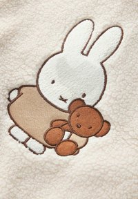 Lapin blanc en robe beige tenant un ours en peluche marron brodé sur un tissu doux couleur crème.