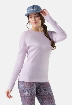 Mujer con trenza larga que lleva una camiseta de manga larga color púrpura pastel, leggings estampados y una gorra azul con texto blanco.