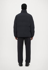 Puffer jacket in textured dark gray with a high collar, boxy silhouette, and elastic cuffs, paired with black pants and shoes.
