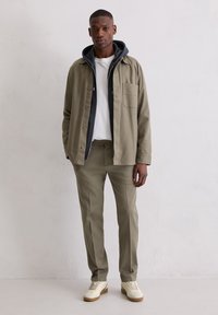 Khakifarbene Jacke über einem marineblauen Hoodie, weißem T-Shirt und passenden Hosen. Verfügt über eine Brusttasche, glatten Stoff und leichte Turnschuhe.