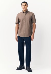 Camisa marrón de manga corta con botones y un bolsillo en el pecho, combinada con vaqueros de pierna recta azul oscuro y zapatos negros con cordones. Fondo liso.