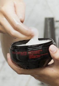Producto capilar Shu Uemura en un tarro redondo negro con texto rojo; textura cremosa blanca siendo recogida con los dedos; capacidad de 200 ml.
