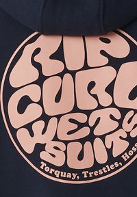 Navy hoodie met een grote, zachte roze cirkelvormige grafiek met gestileerde tekst "CURIOUS WETSUITS" en locatiegegevens onderaan.