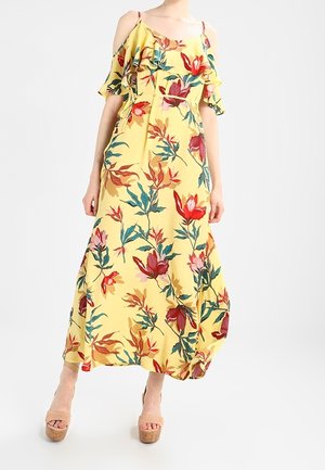 Gul off-shoulder maxikjole med røde og pink blomsterprint, flæsekant ved halsudskæringen og spaghetti-stropper, stylet med beige plateauhøjhælede sko.