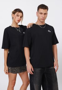 Černá oversize trička z bavlny s bílým hvězdicovým logem na hrudi. Pozadí je čistě bílé. Model pózuje společně.