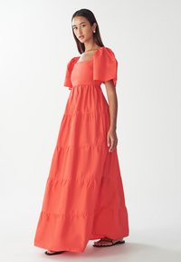 Maksikleita spilgtā korallkrāsā, ar kvadrātu izgriezumu, īsām pūkainām piedurknēm un līmeņu malu dizainu ar plūstošu siluetu.